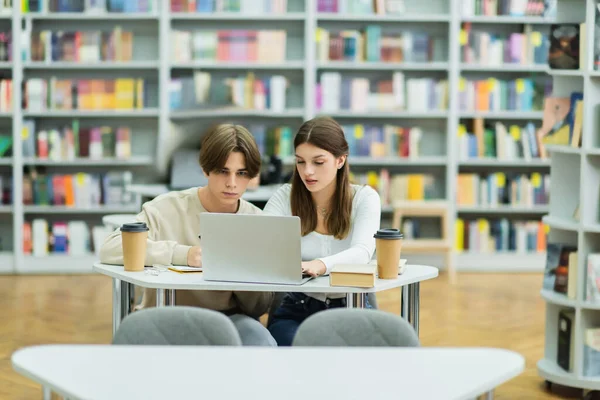 Couple of teenagers studying near laptop and paper cups in library - foto de stock