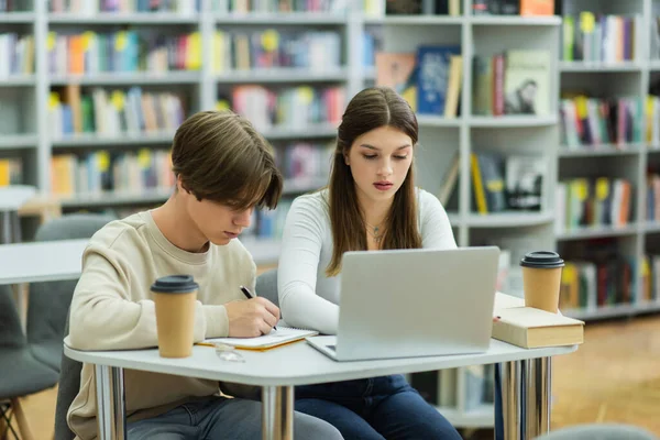 Teenage student writing in notebook near laptop and friend in reading room — Photo de stock