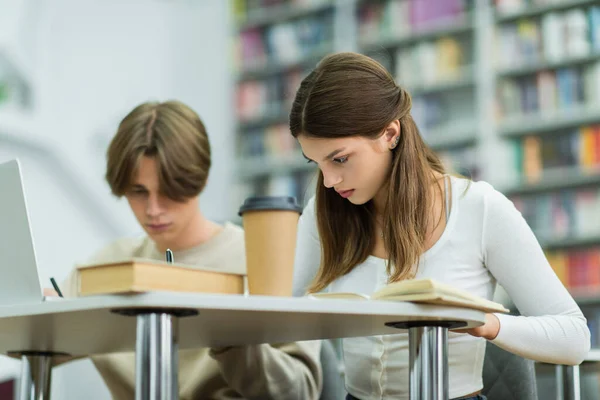 Couple of teenage students writing near laptop while studying in library reading room — Stock Photo