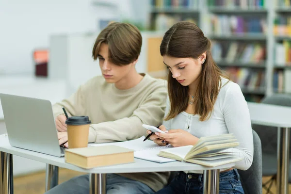 Teenage girl using smartphone near student and laptop in library reading room — Stock Photo