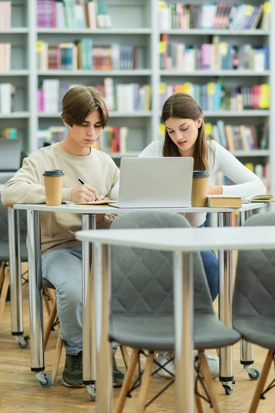 Student in eyeglasses writing near teenage girl and laptop in library — Photo de stock