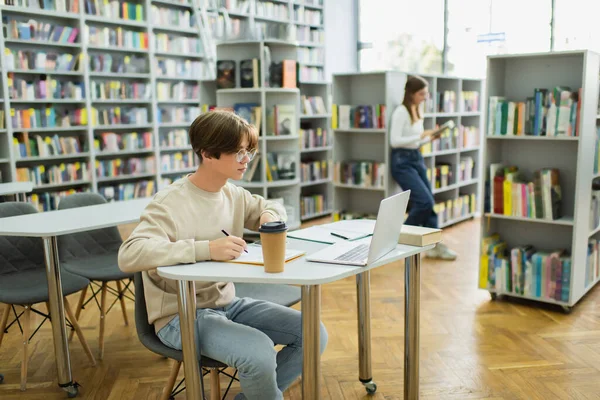 Teen guy in eyeglasses looking at laptop near friend reading book on blurred background - foto de stock