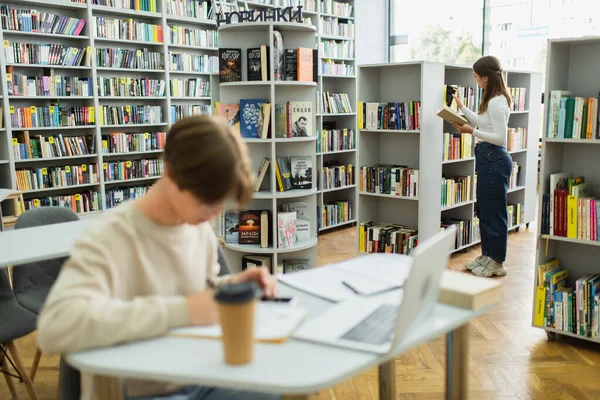Teen girl choosing books while her friend sitting near laptop on blurred foreground - foto de stock