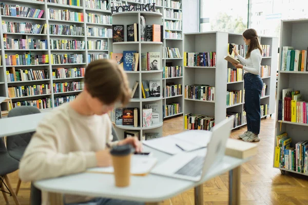 Teen girl choosing books while her friend sitting near laptop on blurred foreground — Stock Photo
