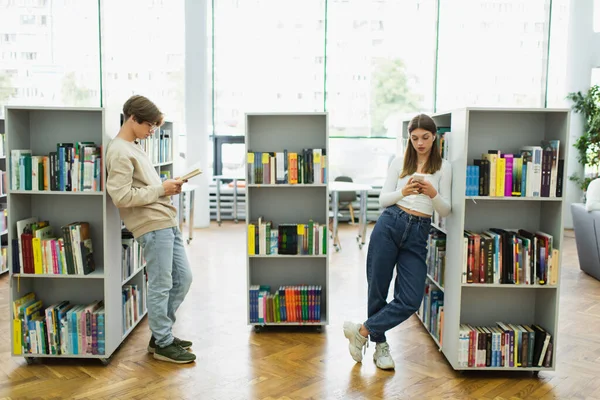 Подростков с книгой и мобильным телефоном, стоящих возле полок в библиотеке — стоковое фото