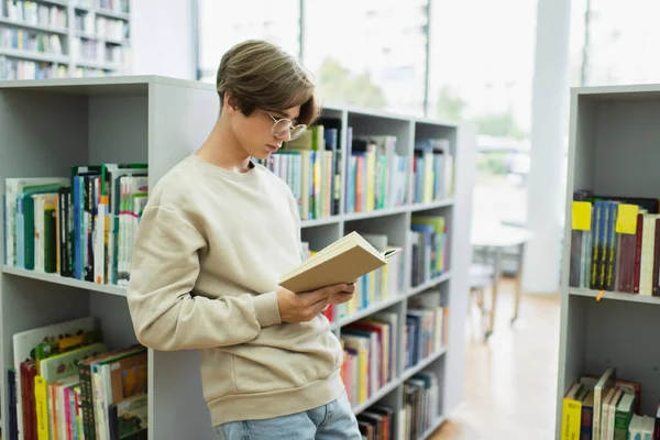 Ученик в очках читает книгу, стоя возле полок в библиотеке — стоковое фото
