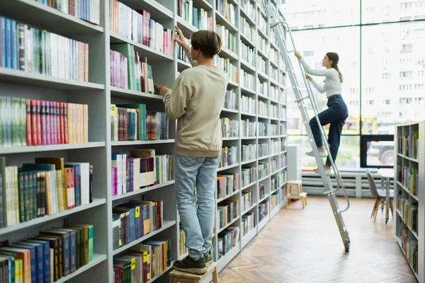Full length of teenage students choosing books on racks in library — Foto stock