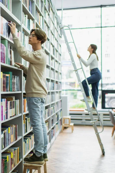 Student in eyeglasses choosing books in library near teenage girl on ladder — Stock Photo