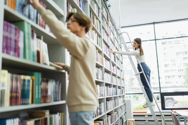 Девочка-подросток, стоящая на лестнице и выбирающая книги рядом с другом на размытом переднем плане — стоковое фото