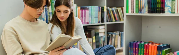 Couple of students reading book while sitting near shelves in library, banner — Stock Photo