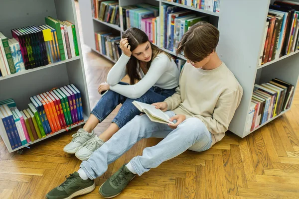 Симпатичная девочка-подросток смотрит на друга, читающего книгу на полу в библиотеке — стоковое фото