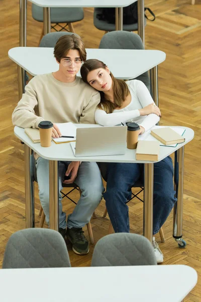 Teenage couple looking at camera near laptop and books in library reading room - foto de stock