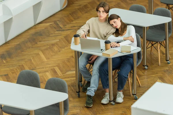 Teenage students looking at camera while sitting in reading room near laptop and paper cups - foto de stock