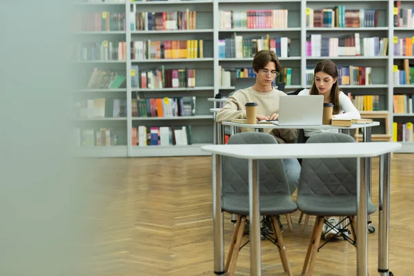 Couple of teenagers sitting near laptop and paper cups in library on blurred foreground — Stock Photo
