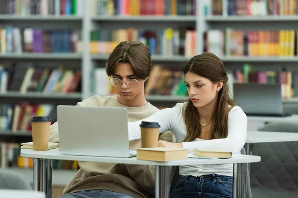 Teenage girl and guy in eyeglasses studying near laptop and books in library - foto de stock