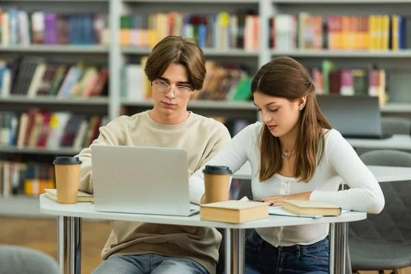 Teenage students studying near laptop and paper cups in blurred library — Stock Photo