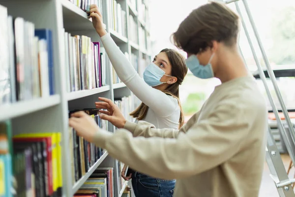 Девушка в медицинской маске выбирает книги в библиотеке рядом с другом на размытом переднем плане — стоковое фото