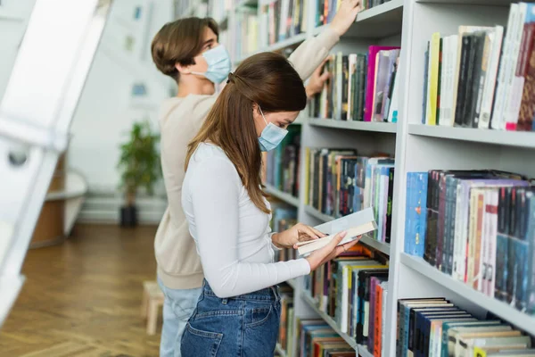 Side view of teenage girl in medical mask reading book near rack and guy on blurred background — Stock Photo