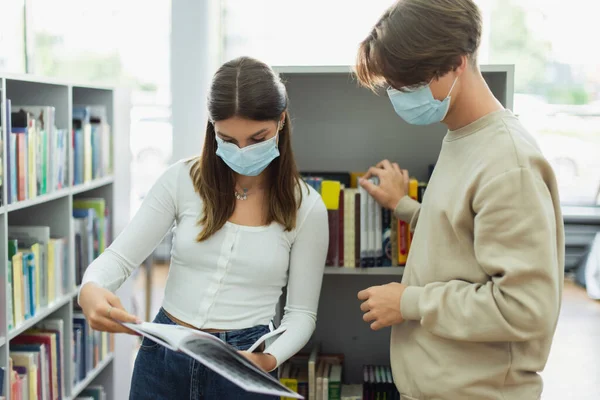 Teenage girl in medical mask showing book to guy near racks in library — Stock Photo
