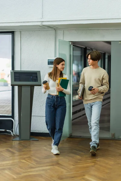 Full length of teenage friends with coffee to go and notebooks entering library — Stock Photo