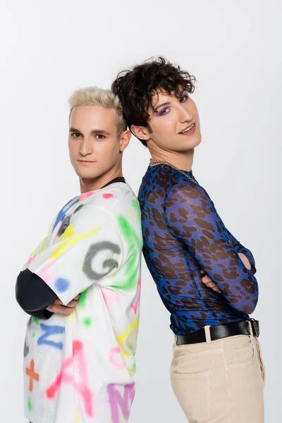 Blonde gay man and smiling queer person standing back to back with crossed arms isolated on grey - foto de stock