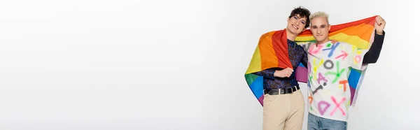 Trendy queer person and blonde gay man with lgbtq flag smiling at camera isolated on grey, banner — Stock Photo