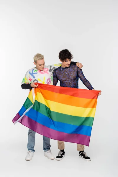Blonde gay man and brunette nonbinary person standing with lgbtq flag on grey background - foto de stock