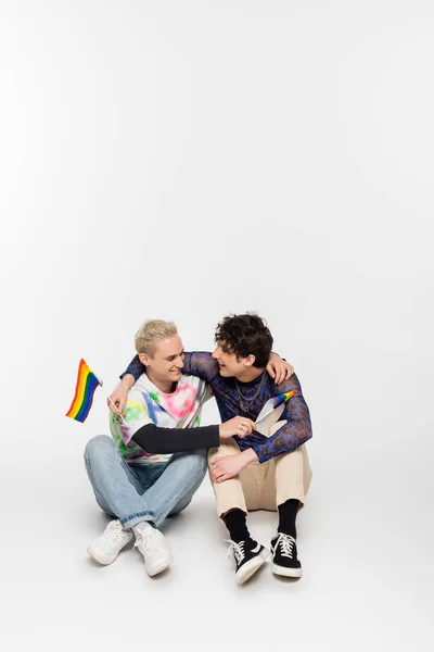 Happy gay man and nonbinary person with small lgbtq flags embracing while sitting on grey background — стоковое фото