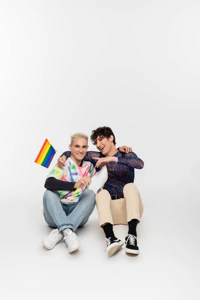 Усміхнений гей-чоловік з маленькими прапорами lgbtq, що сидять поруч з квір-особою, вказуючи пальцем на сірому фоні — стокове фото