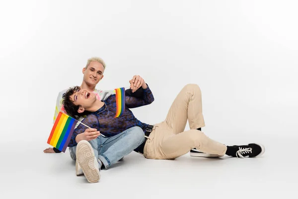 Queer person with small lgbtq flags laughing near gay man while sitting on grey background - foto de stock