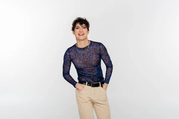 Smiling nonbinary person standing with hands in pockets of beige trousers isolated on grey - foto de stock