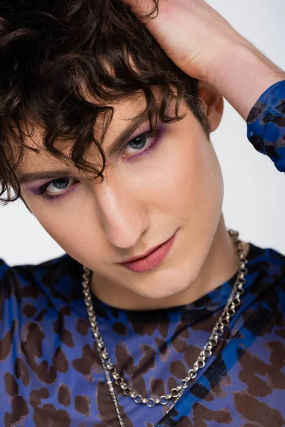 Portrait of nonbinary person with makeup and silver necklaces touching wavy hair isolated on grey — Stock Photo