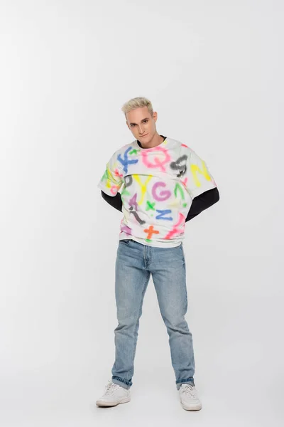 Full length of gay man in jeans and t-shirt with alphabet print posing with hands behind back on grey background - foto de stock
