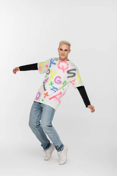 Full length of gay in alphabet print t-shirt and jeans posing on tiptoes on grey background — Stock Photo