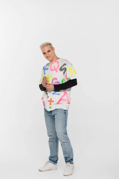 Full length of blonde fashionable gay man smiling at camera on grey background - foto de stock