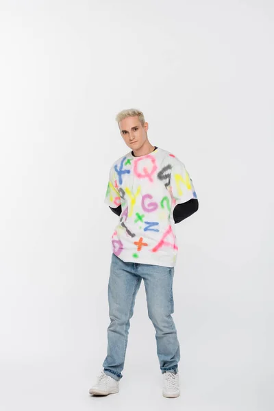 Full length of young gay man in jeans and t-shirt with multicolored alphabet print on grey background — Stockfoto