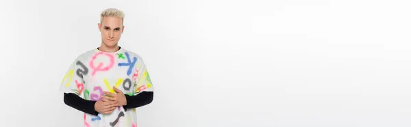 Молода блондинка гей в стильній різнокольоровій футболці дивиться на камеру ізольовано на сірому, банер — стокове фото
