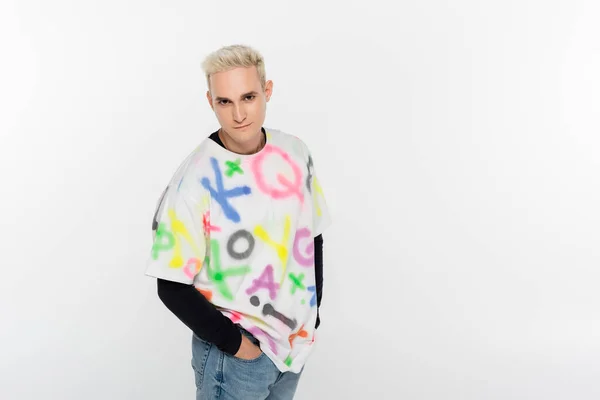 Young gay man standing with hands in pockets and looking at camera isolated on grey — Stock Photo