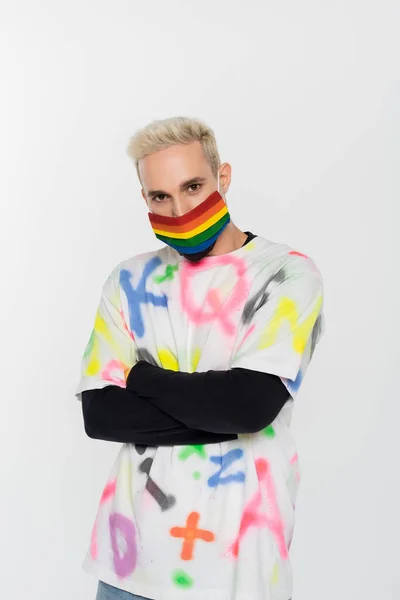 Young gay man in rainbow colors medical mask standing with crossed arms isolated on grey — Stockfoto