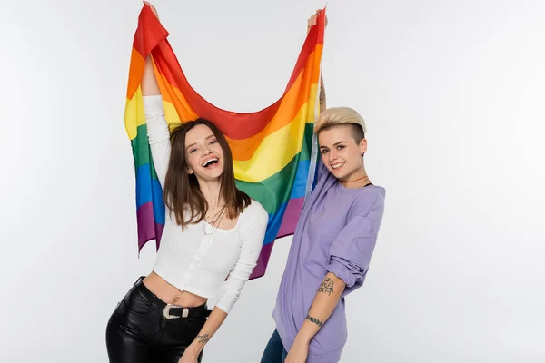 Excited same sex couple holding lgbtq flag and smiling at camera isolated on grey - foto de stock