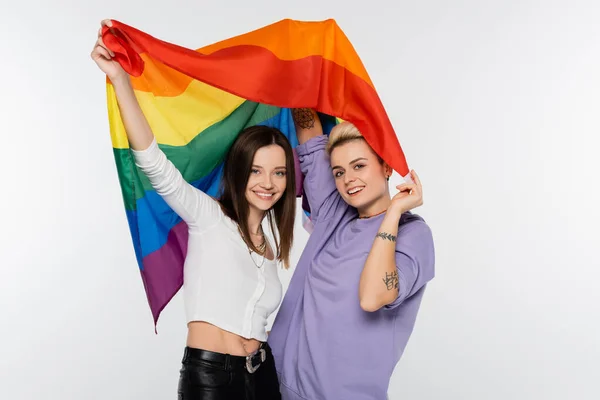 Happy and trendy lesbians looking at camera while holding lgbtq flag isolated on grey - foto de stock