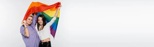 Young and joyful lesbians with lgbtq flag looking at camera isolated on grey, banner — Photo de stock