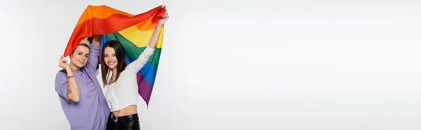 Молоді та радісні лесбійки з прапором lgbtq дивиться на камеру ізольовано на сірому, банер — стокове фото