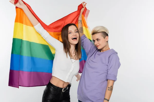 Tattooed lesbian woman with excited girlfriend holding lgbtq flag isolated on grey — Foto stock