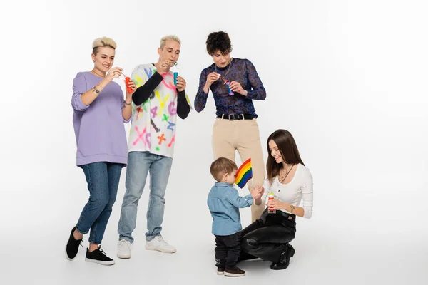 Young lgbtq community friends blowing soap bubbles near toddler boy on grey background — стоковое фото