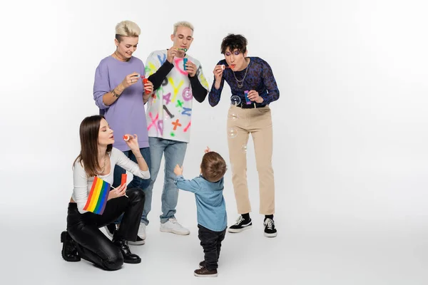 Lesbian woman with rainbow flag blowing soap bubbles near toddler boy and lgbtq community friends on grey — Stockfoto