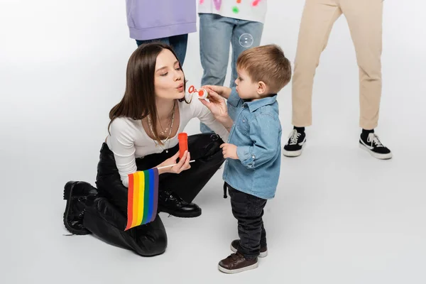 Young lesbian woman with lgbtq flag blowing soap bubbles near toddler boy on grey background — Fotografia de Stock