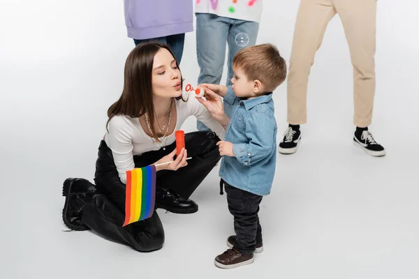 Young lesbian woman with lgbtq flag blowing soap bubbles near toddler boy on grey background — Stock Photo