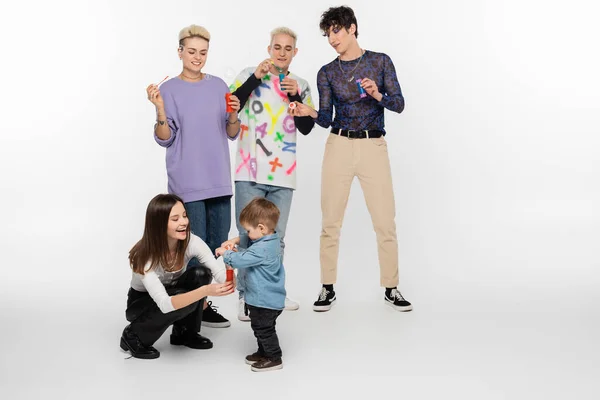 Happy lgbtq friends blowing soap bubbles near toddler boy on grey background — Stockfoto