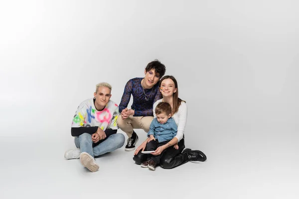 Happy lesbian woman sitting with toddler boy near gay man and queer person on grey background - foto de stock
